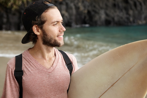 Hobby, vrije tijd en avontuur. Jonge surfer met schattige glimlach die zijn surfplank onder zijn arm draagt en oceaan bekijkt, die gelukkige uitdrukking heeft