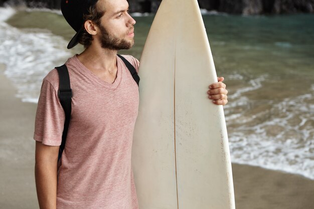Hobby, watersport en actieve levensstijl concept. Jonge surfer die honkbal GLB dragen die zich achteruit alleen op zandig strand bevinden en op zee met nadenkende uitdrukking kijken