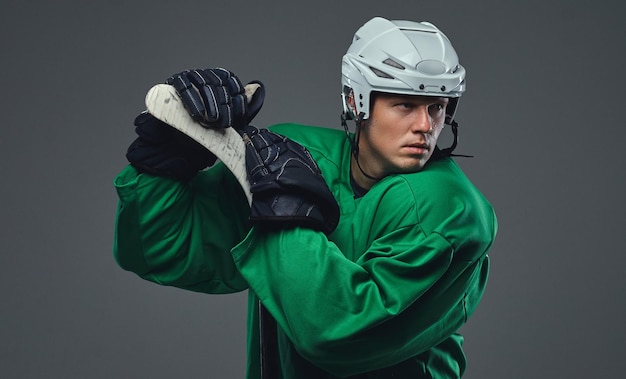 Hockeyspeler die groene beschermende kleding en witte helm draagt die zich met de hockeystick op een grijze achtergrond bevinden.