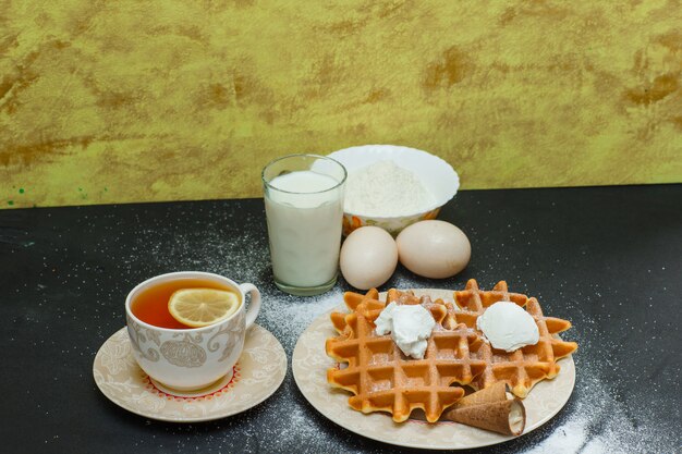 Hoge hoek bekijken wafels in plaat met thee, eieren, meel op donkere en gestructureerde oppervlak. horizontaal