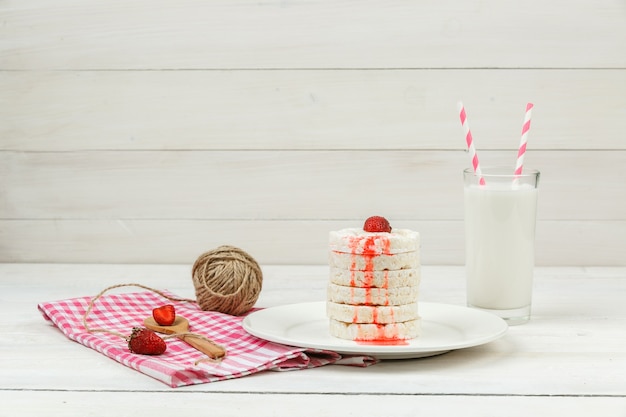 Hoge hoek bekijken witte rijstwafels op plaat met aardbeien, schuimgebak, kluwen van touw en melk op witte houten plank oppervlak.