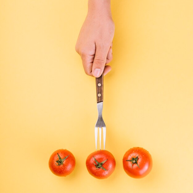 Hoge hoekmening van de hand die van een persoon vork in rode tomaat op gele achtergrond opneemt