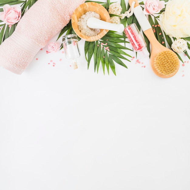 Gratis foto hoge hoekmening van handdoek; zout; loofah; verlaat; fles; borstel en nep bloemen op wit oppervlak