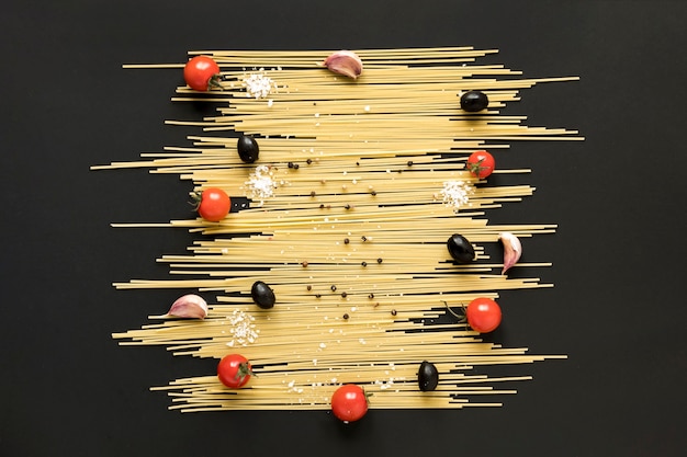 Hoge hoekmening van ruwe spaghettideegwaren; kerstomaat; zwarte olijven en zwarte peper gerangschikt op zwart oppervlak