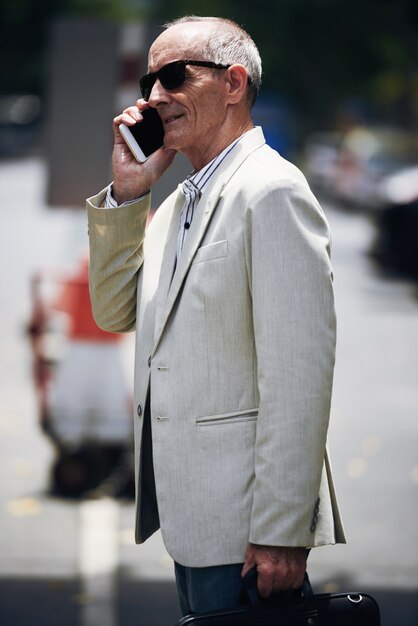 Hogere Kaukasische zakenman in zonnebril die zich in straat bevindt en op telefoon spreekt