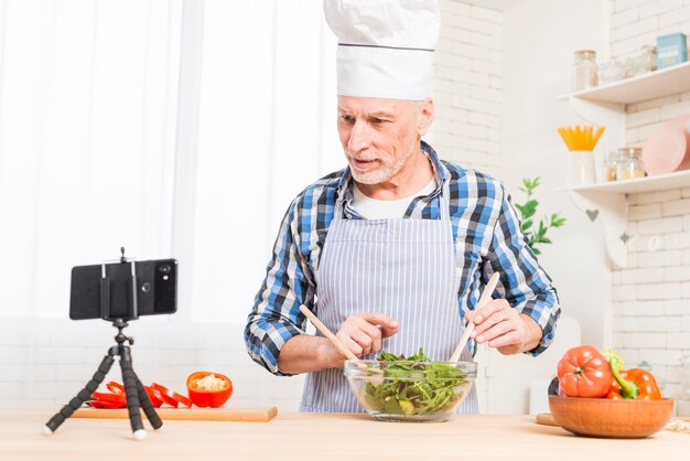 Hogere mens die mobiele telefoon bekijken terwijl het voorbereiden van de salade in de keuken