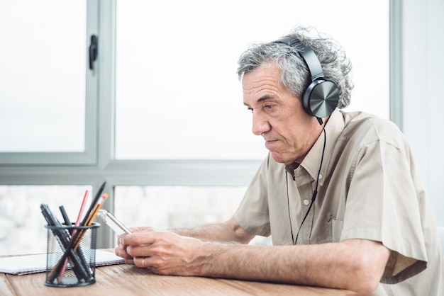 Hogere mens het luisteren muziek op hoofdtelefoon bij bureau dichtbij het venster