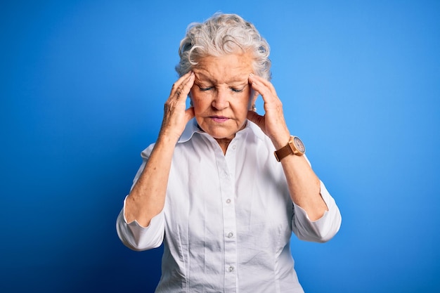 Gratis foto hogere mooie vrouw, gekleed in een elegant shirt dat over een geïsoleerde blauwe achtergrond staat met de hand op het hoofd voor pijn in het hoofd omdat stress lijdt aan migraine