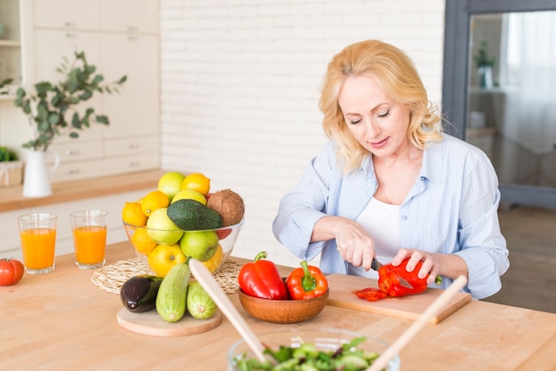 Hogere vrouw die de rode groene paprika met mes op houten lijst snijdt