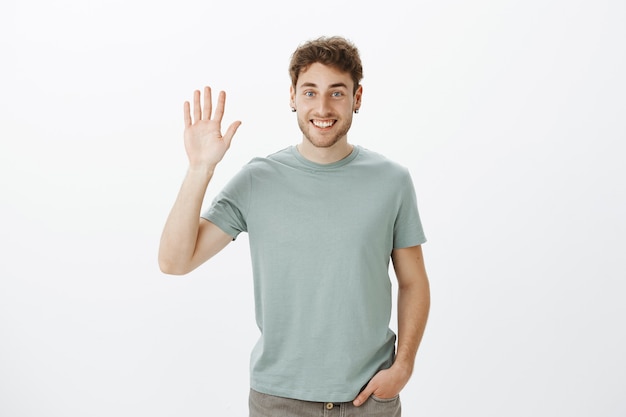 Hoi leuk je te ontmoeten. Portret van knappe uitgaande Europese man in casual t-shirt hand opheffen en palm in hallo gebaar zwaaien