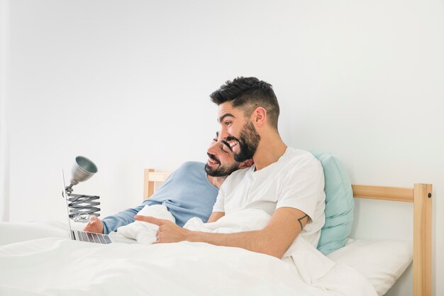 Homoseksueel paar die op bed liggen die terwijl het bekijken laptop tegen witte muur lachen