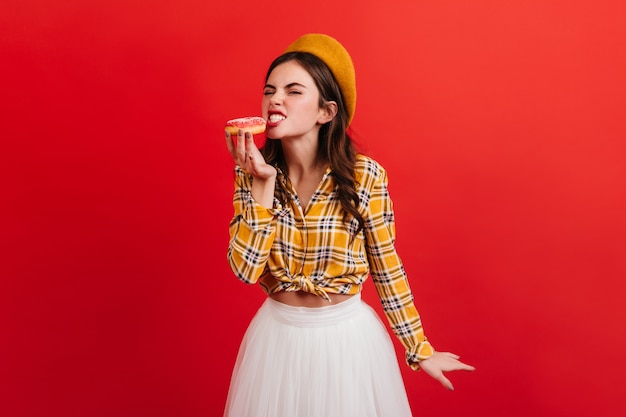 Hongerig meisje in stijlvolle outfit bijt aardbeidoughnut. Portret van vrouw in geruite blouse en oranje baret op lichte muur.