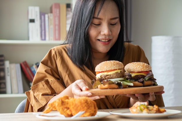 Hongerige vrouw met overgewicht die hamburger vasthoudt op een houten bord fried chicken en pizza op tafel