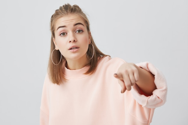 Gratis foto hoofd en schouders van jonge charmante blanke vrouw, gekleed in roze stijlvolle sweatshirt met recht blond haar in evenwicht tegen de grijze blinde muur op zoek en wijst naar je