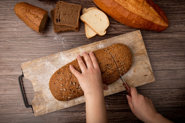 Hoogste mening die van vrouwenhanden sandwichbrood op scherpe raad en ander brood op houten achtergrond snijden