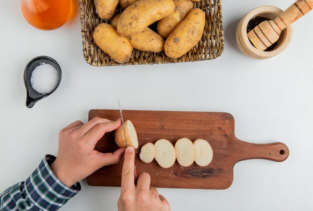 Hoogste mening van handen die aardappel met mes op scherpe raad snijden en andere in plaat boter zoute zwarte peper op witte oppervlakte