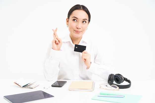 Hoopvolle aziatische kantoormedewerker toont creditcard en gekruiste vingers voor geluk maakt wens wit