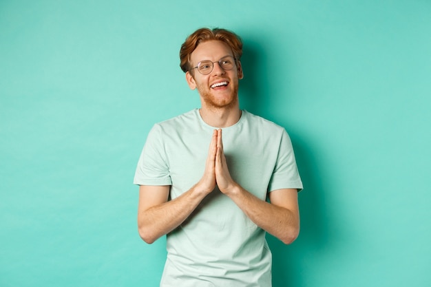 Hoopvolle roodharige man met baard, bril en t-shirt dragend, hand in hand in namaste of smekend gebaar en naar rechts kijkend, glimlachend en bedankend, staande over turkooizen achtergrond