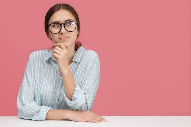 Horizontaal beeld van doordachte mooie jonge vrouw die bril draagt die weg kijkt met peinzende dromerige glimlach, hand op kin houdt, bedrijfsstrategie ontwikkelt, veel creatieve ideeën heeft