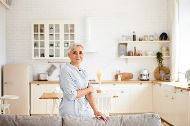 Horizontaal beeld van prachtige vrolijke bejaarde zestigjarige huisvrouw die rust heeft na het schoonmaken van alle kamers, met vrolijke gelaatsuitdrukking, staande in de woonkamer met keuken in