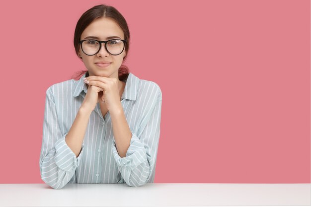 Horizontaal beeld van vrolijk zelfverzekerd schattig jong wijfje dat gestreept overhemd en een bril draagt die handen onder haar kin plaatst, die bij wit bureau werkt. Baan, beroep en werkgelegenheid concept