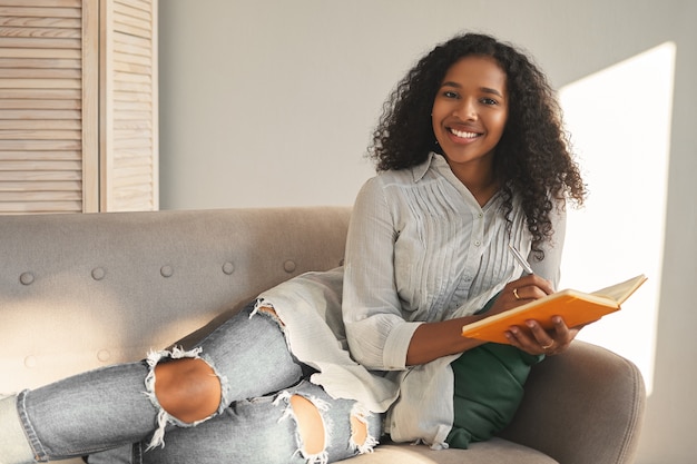 Horizontaal portret van charmante positieve jonge gemengd ras vrouw in stijlvol shirt en gescheurde spijkerbroek breed glimlachend, liggend op de bank met dagboek, vakantie plannen, gelukkig en geïnspireerd voelen