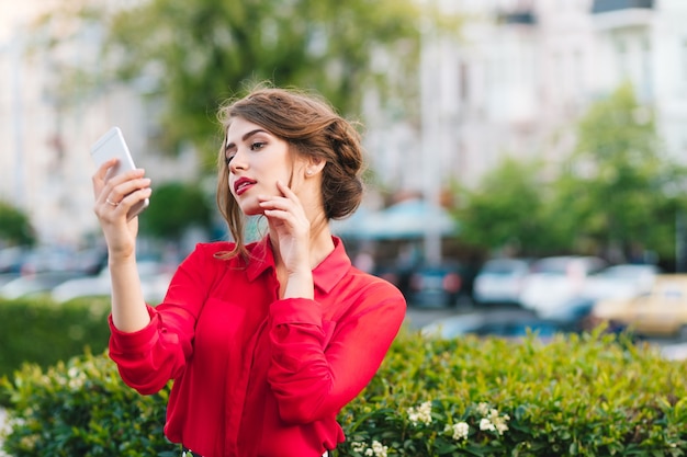 Horizontaal portret van mooi meisje dat zich in park bevindt. Ze draagt een rode blouse en een mooi kapsel. Ze kijkt naar de telefoon in de hand en droomt.