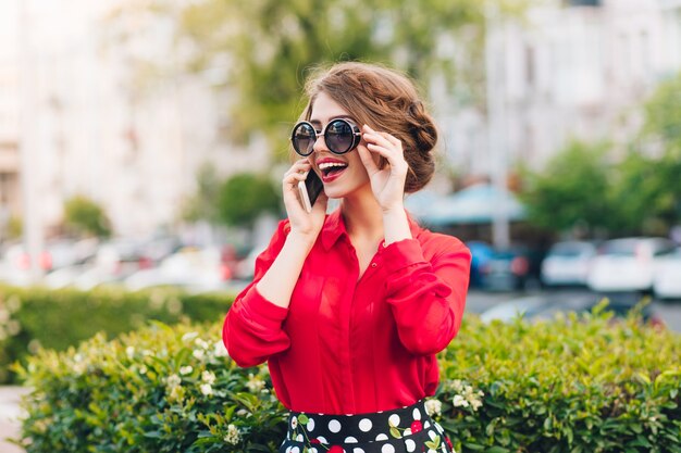 Horizontaal portret van mooi meisje die in zonnebril in park lopen. Ze draagt een rode blouse en een mooi kapsel. Ze is aan het bellen.