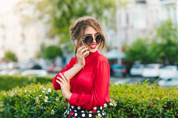 Horizontaal portret van mooi meisje in zonnebril die zich in park bevinden. Ze draagt een rode blouse en een mooi kapsel. Ze is aan het bellen.