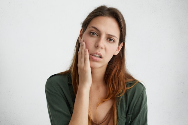 Horizontaal portret van vrij droevige vrouw met heldere donkere ogen en steil haar die hand op wang houden die boos kijken