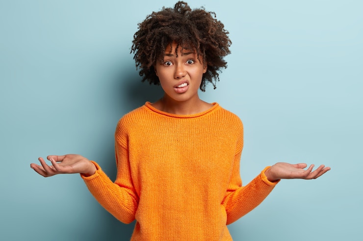 Gratis Foto horizontaal schot van aantrekkelijke vrouw met verbaasde uitdrukking, spreidt handen met verwarde blik, fronst gezicht, draagt oranje kleren, voelt zich aarzelend, verontwaardigd geïsoleerd over blauwe muur. twijfel concept