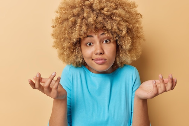 Horizontaal schot van aarzelende vrouw met krullend haar haalt schouders op kijkt met verbijstering voelt zich verward draagt casual blauwe trui geïsoleerd over bruine achtergrond Clueless meisje gebaren in verwarring