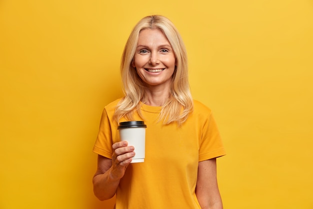 Horizontaal schot van blonde Europese vrouw met aangename glimlach minimale make-up houdt wegwerp kopje koffie gekleed in casual t-shirt