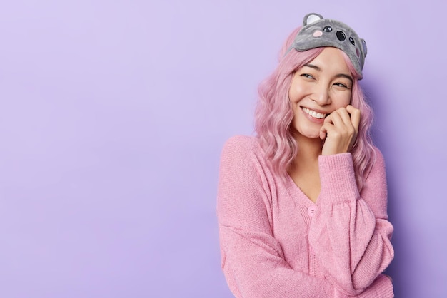Horizontaal schot van dromerige aziatische vrouw raakt gezicht met handen heeft tedere uitdrukking glimlacht vrolijk draagt casual trui en slaapmasker geïsoleerd over puple achtergrond lege kopie ruimte. oprechte emoties