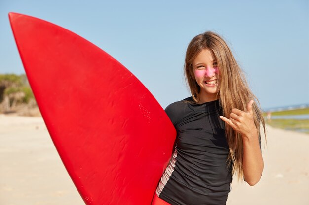 Horizontaal schot van gelukkig meisje geniet van goede weersomstandigheden om te surfen, maakt shaka of hangt los gebaar