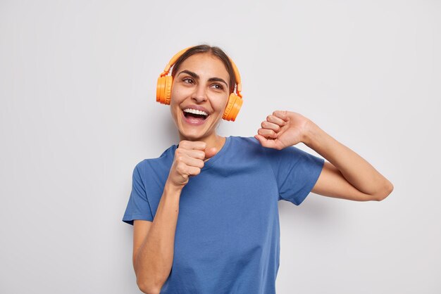 Horizontaal schot van gelukkige brunette jonge vrouw houdt hand in de buurt van mond zingt favoriete liedje draagt casual blauwe t-shirt beweegt met ritme van muziek poses tegen witte achtergrond geniet van nieuwste afspeellijst