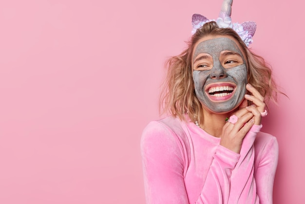Horizontaal schot van gelukkige jonge Europese vrouw met voedend gezichtsmasker ondergaat huidverzorgingsprocedures kijkt weg glimlacht breed poseert tegen roze achtergrond met kopieerruimte voor uw promo