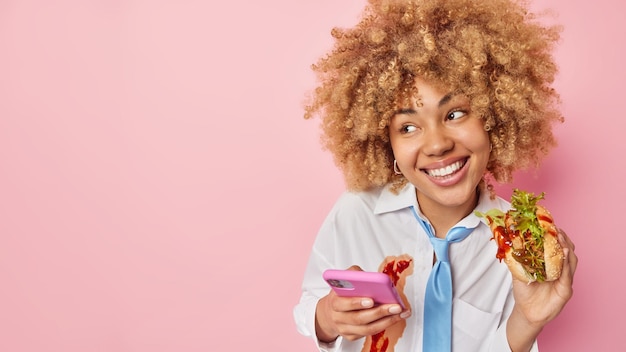 Horizontaal schot van gelukkige jonge vrouw met krullend haar heeft een smakelijke snack geniet van het eten van heerlijke hamburger lijdt aan ongezonde gewoonte om te veel te eten houdt mobiele telefoon draagt wit overhemd en blauwe stropdas