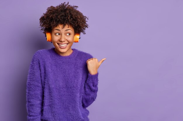 Horizontaal schot van goed uitziende vrolijke Afro-Amerikaanse vrouw luistert audiotrack wijst duim weg op lege ruimte glimlach positief poses