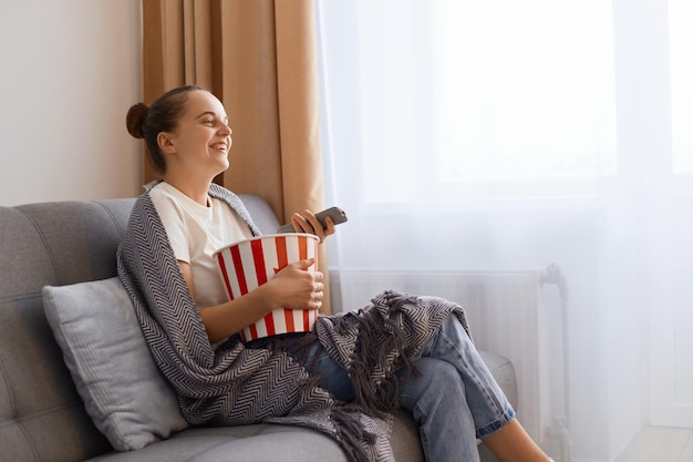 Horizontaal schot van lachende gelukkige positieve vrouw zittend op de bank gewikkeld in een deken en tv kijken grappige comedyshow uiting van geluk genieten van tijd doorbrengen alleen thuis Premium Foto