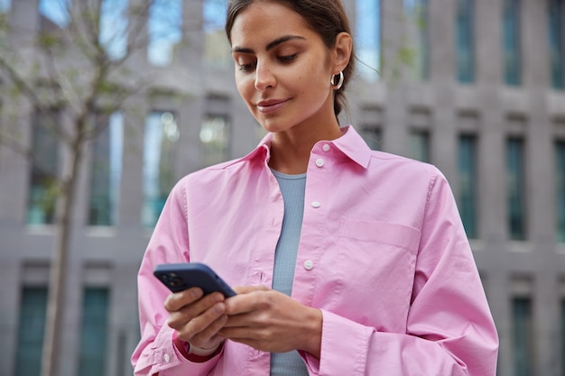 Horizontaal schot van mooi millennial meisje gebruikt mobiele telefoon in de stad voor het vinden van route verkent nieuwe bezienswaardigheden draagt roze shirt poses op wazig gebouw