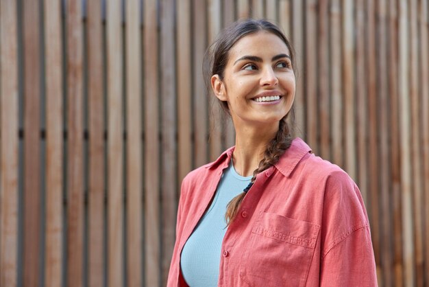 Horizontaal schot van mooie glimlachende vrouw met donker haar gekamd in vlecht gekleed in een casual roze shirt, weggefocust met blije uitdrukking die overdag naar buiten wandelt tegen houten vandaar de achtergrond