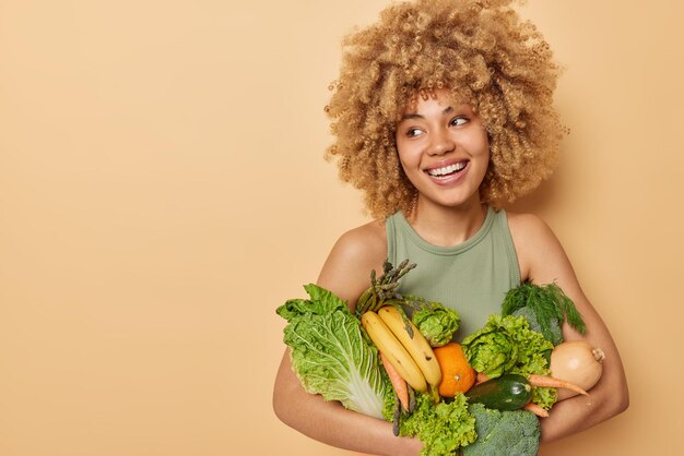 Horizontaal schot van mooie vrouw draagt een verscheidenheid aan verse groenten en fruit consumeert kruidenierswinkel vol vitamines kijkt weg staat tegen beige achtergrond lege ruimte voor uw promotionele inhoud