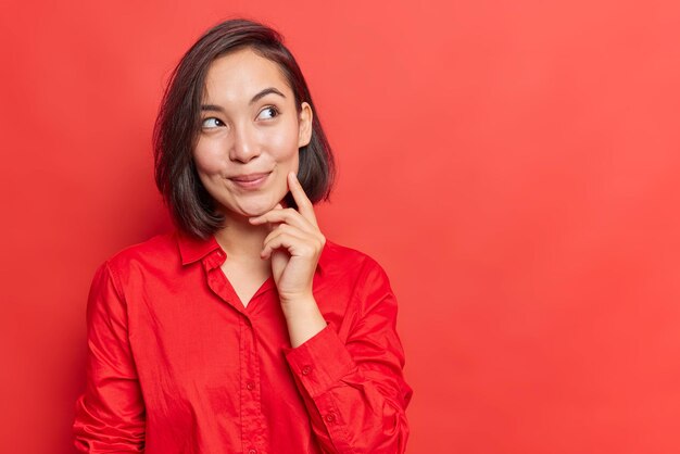 Horizontaal schot van mooie vrouw met donker haar houdt vinger op wang kijkt weg heeft dromerige uitdrukking draagt shirt geïsoleerd over rode achtergrond blanco kopieerruimte voor uw promotionele inhoud