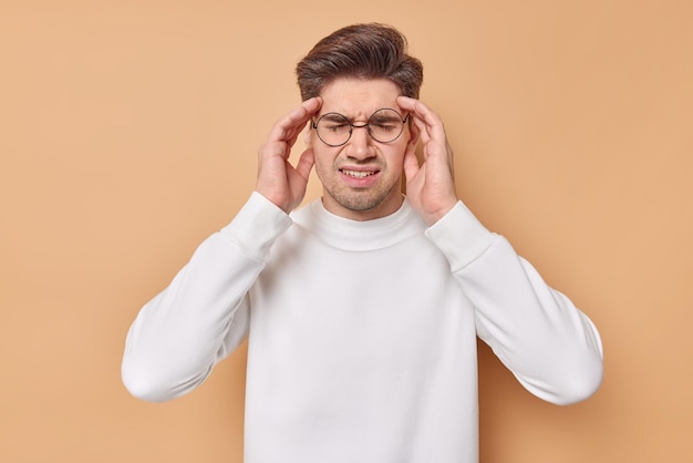 Gratis foto horizontaal schot van ontevreden jongeman lijdt aan migraine houdt handen op slapen heeft ernstige hoofdpijn probeert pijn te onthullen draagt bril en witte trui geïsoleerd over beige achtergrond