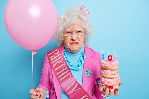 Horizontaal schot van ontevreden senior vrouw fronst gezicht heeft ongelukkige stemming houdt stapel smakelijke geglazuurde donuts met brandende kaars opgeblazen ballon