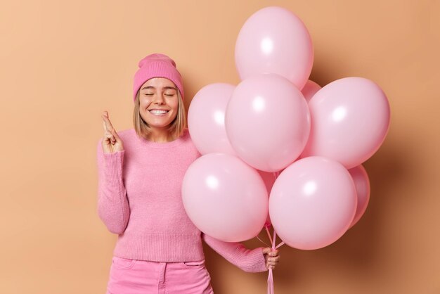 Horizontaal schot van positieve jonge glimlachende vrouw doet een wens op verjaardag houdt vinger gekruist glimlacht breed hoopt dat dromen uitkomen draagt roze hoed trui en broek geïsoleerd over bruine achtergrond