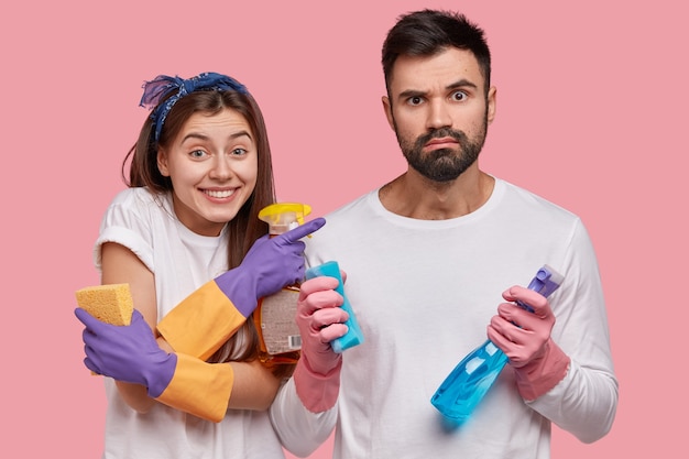 Horizontaal schot van positieve jonge vrouw wijst naar echtgenoot die geërgerde uitdrukking heeft, schoon huis samen, niet van vuil houdt