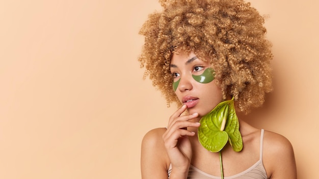 Horizontaal schot van tedere vrouw met krullend borstelig haar houdt de hand dichtbij de mond gefocust weg brengt hydrogelpleisters onder de ogen aan om wallen te verminderen houdt calla-bloemhoudingen tegen beige studiomuur