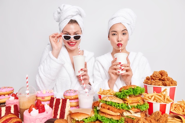 Gratis foto horizontaal schot van twee vrouwen die koolzuurhoudende drank drinken met rietjes en een comfortabele kamerjas dragen